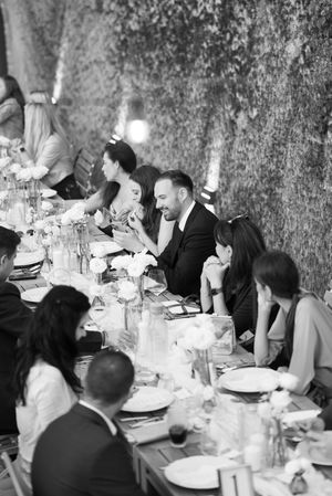 Black and white photo of wedding guests laughing and enjoying dinner at a long decorated reception table.