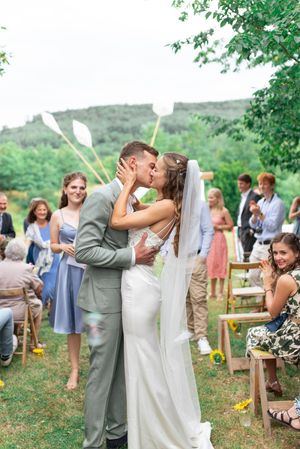 Newlyweds share first kiss at outdoor wedding ceremony surrounded by cheering guests.