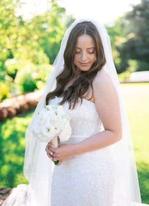Elegant bride in strapless lace gown and cathedral veil holds white rose bouquet in sunlit garden.