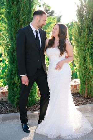 Bride and groom smiling at each other beside tall cypress trees on wedding day.