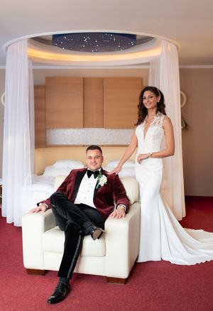 Bride standing beside groom seated in white chair beneath a star-ceiling canopy bed in an elegant hotel room.