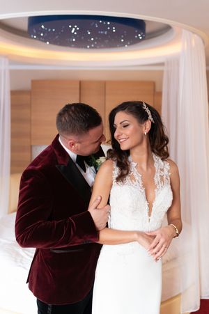 Bride and groom sharing an intimate moment in a luxury hotel suite, groom in burgundy velvet tuxedo embracing smiling bride.