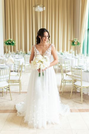 Bride in floral applique tulle wedding gown holding white bouquet in elegant gold and white reception hall.