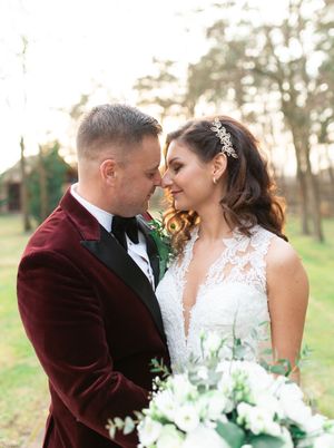 Bride and groom in velvet burgundy tuxedo share intimate moment with white floral bouquet.