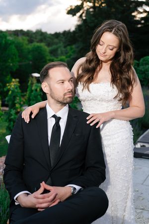 Groom in black suit seated as bride in white textured gown stands behind him outdoors.