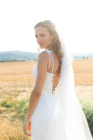 Bride in backless lace wedding gown with veil poses in golden wheat field at sunset.