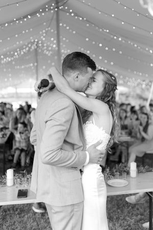 Black and white photo of bride and groom sharing first kiss under a tented reception with fairy lights.