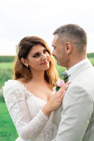 Bride in long-sleeve lace gown gazes lovingly at groom in light suit with pink boutonniere in an outdoor setting.