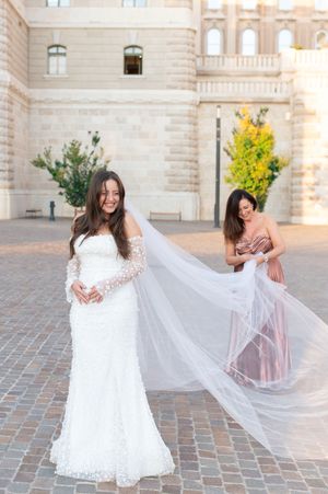 Smiling bride in lace mermaid gown with cathedral veil held by bridesmaid in pink dress.