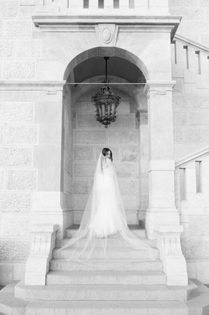 Black and white bridal portrait of bride in cathedral veil standing under ornate stone archway.