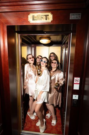 Bride and bridesmaids in satin robes and sunglasses pose together inside a vintage wooden hotel elevator labeled LIFT.