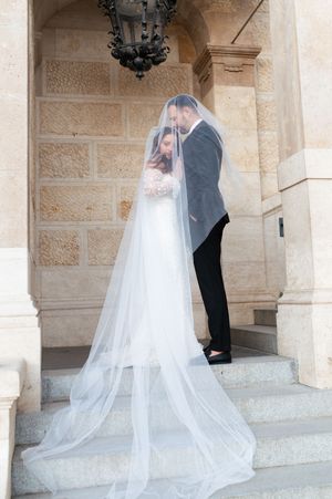 Bride in lace gown with cathedral veil embraces groom on stone steps of ornate building.