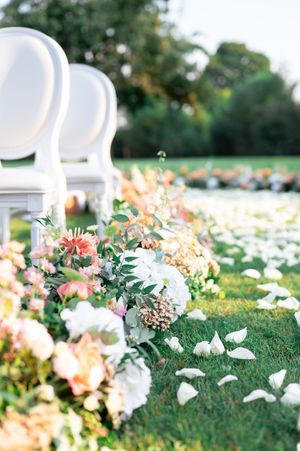Elegant outdoor wedding aisle lined with white chairs and lush floral arrangements of pink and white blooms on green grass.