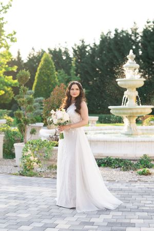 Elegant bride in flowing white gown holding bouquet stands before ornate garden fountain.