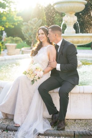 Bride and groom sitting by an ornate stone fountain in golden sunlight, bride holding a pink bridal bouquet.