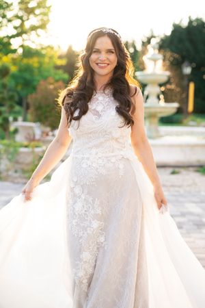 Radiant bride in lace wedding gown with floral appliques smiling joyfully in a sunlit garden with fountain.