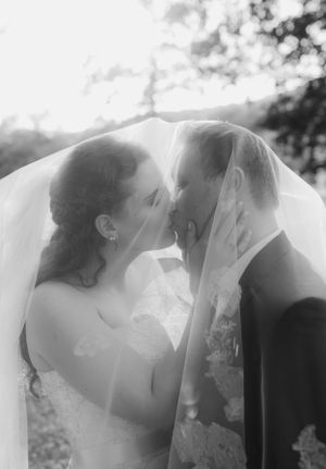 Black and white romantic photo of bride and groom kissing under a sheer wedding veil outdoors.