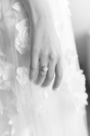 Black and white close-up of bride's hand wearing a delicate diamond ring on her wedding dress.