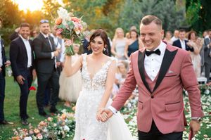 Joyful bride and groom walk down outdoor aisle, bride holding bouquet, groom in pink tuxedo, guests celebrating behind them.