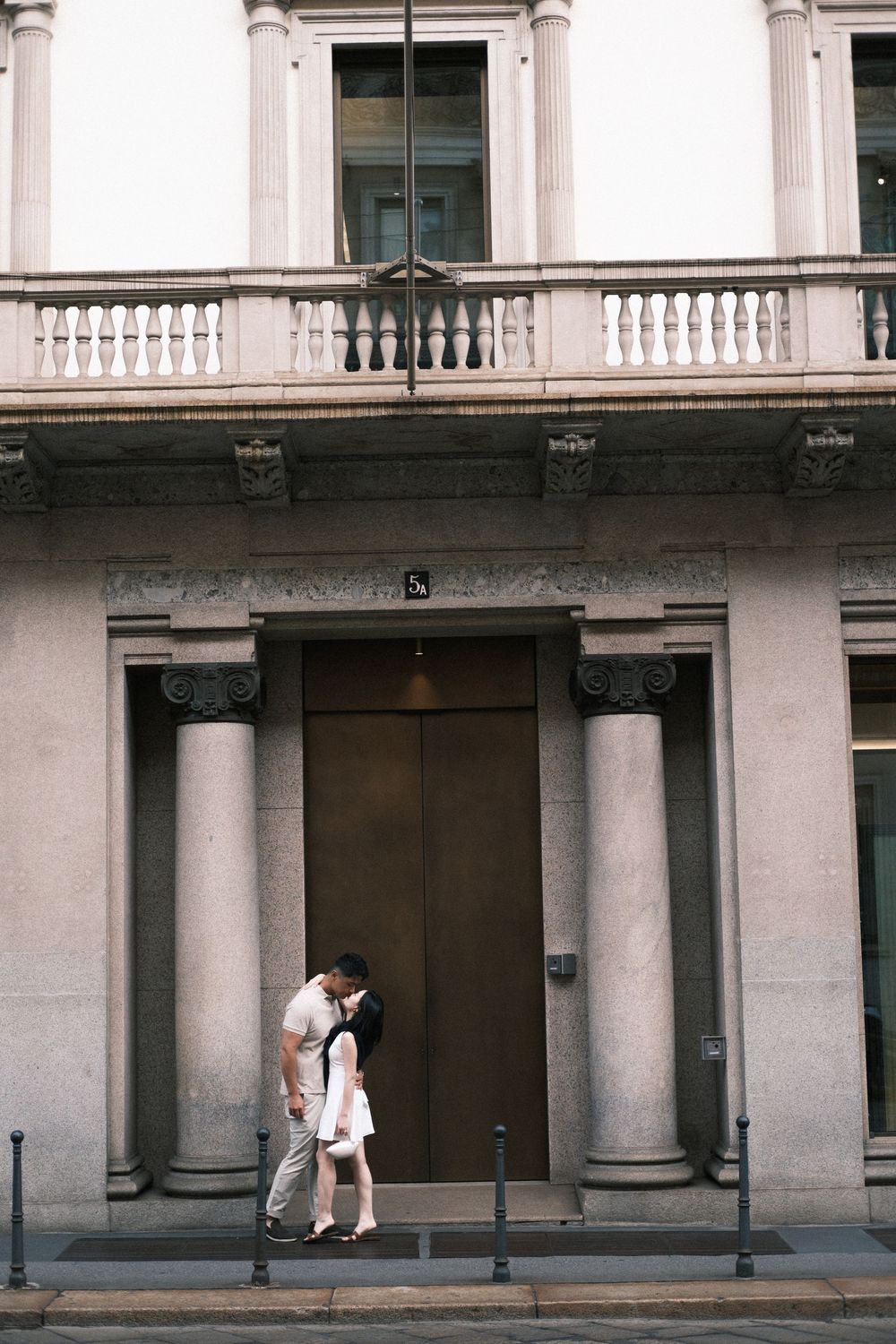 Couple sharing a romantic kiss in front of a grand neoclassical building with tall columns and ornate balustrade.