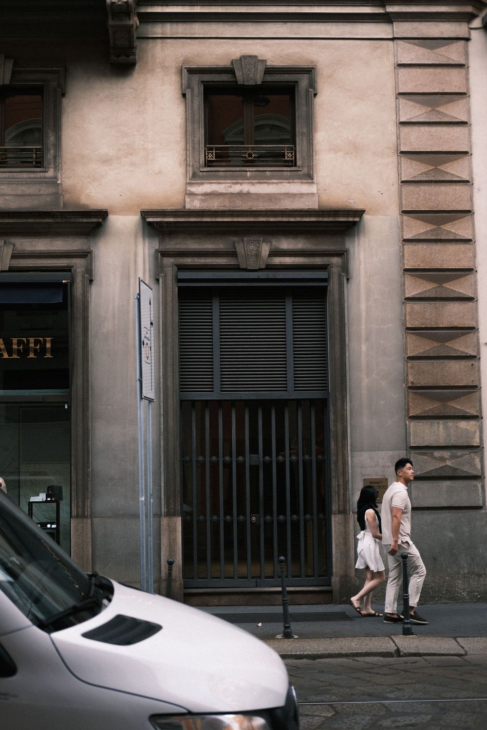Two people walking past a historic stone building with shuttered doors and windows on a European city street.