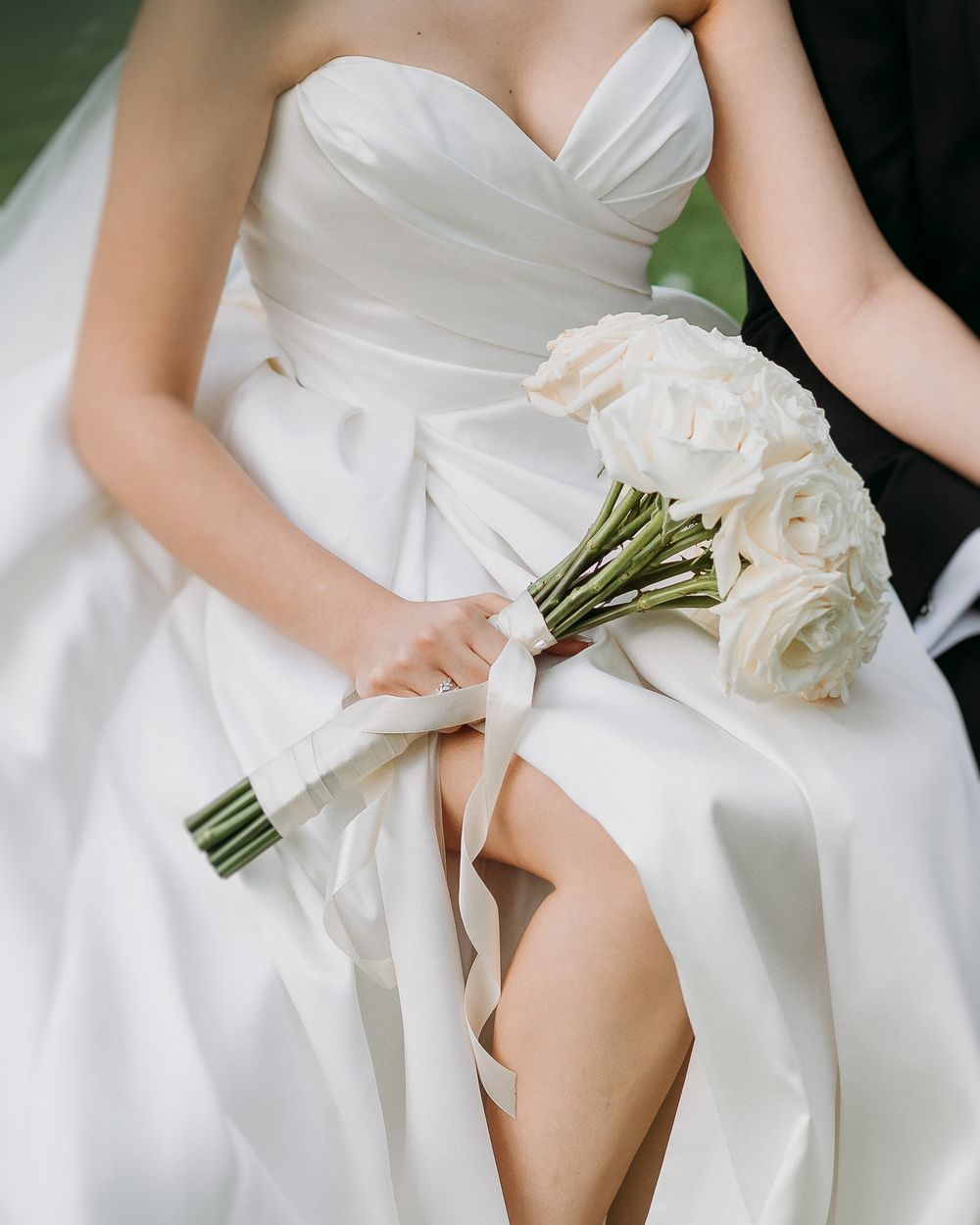 Hand holding open ring box with diamond ring, baby's breath flowers and green gift bag on wooden surface.