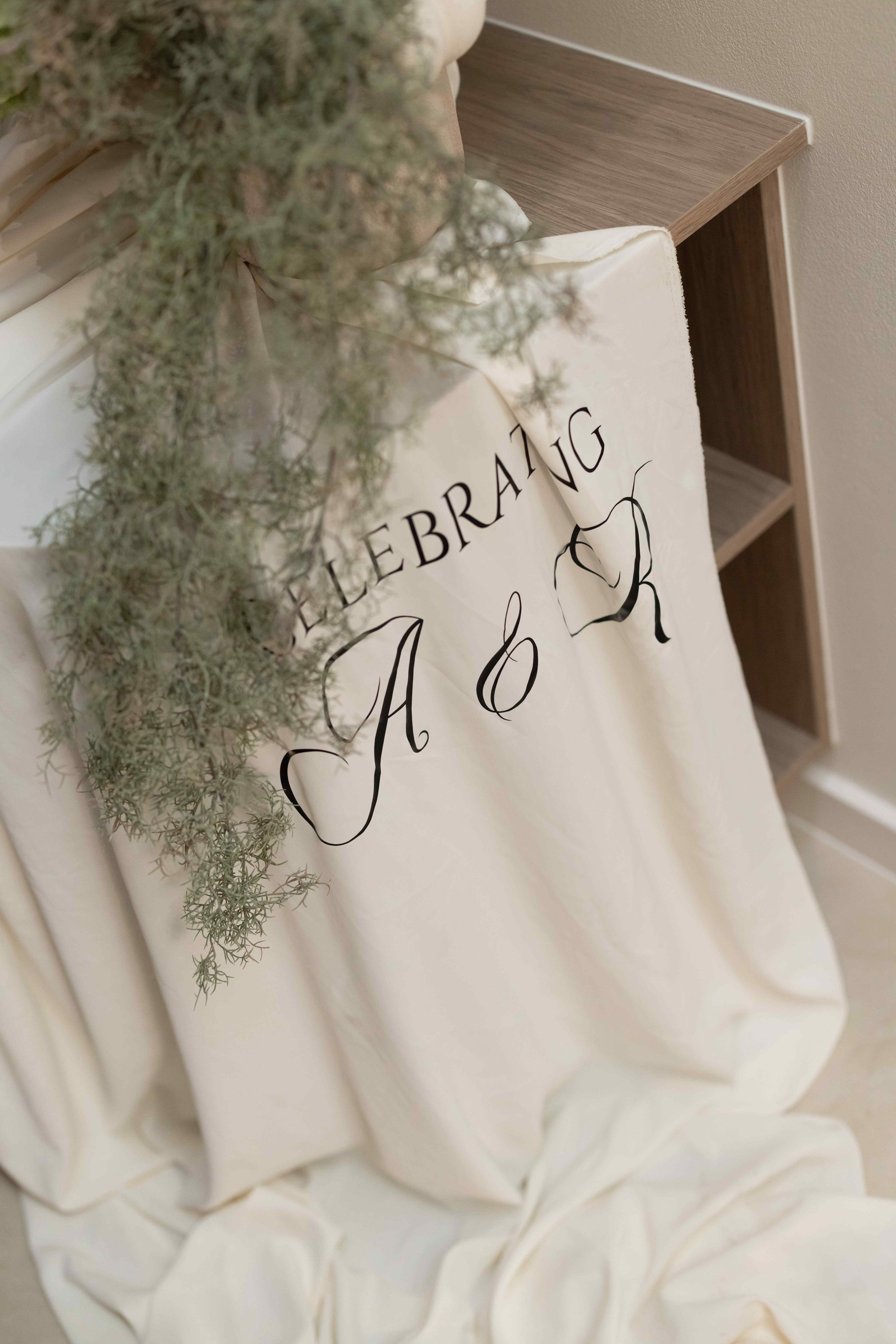 Elegant cream fabric draped over a shelf with calligraphy monogram A&R celebrating wedding signage and greenery.