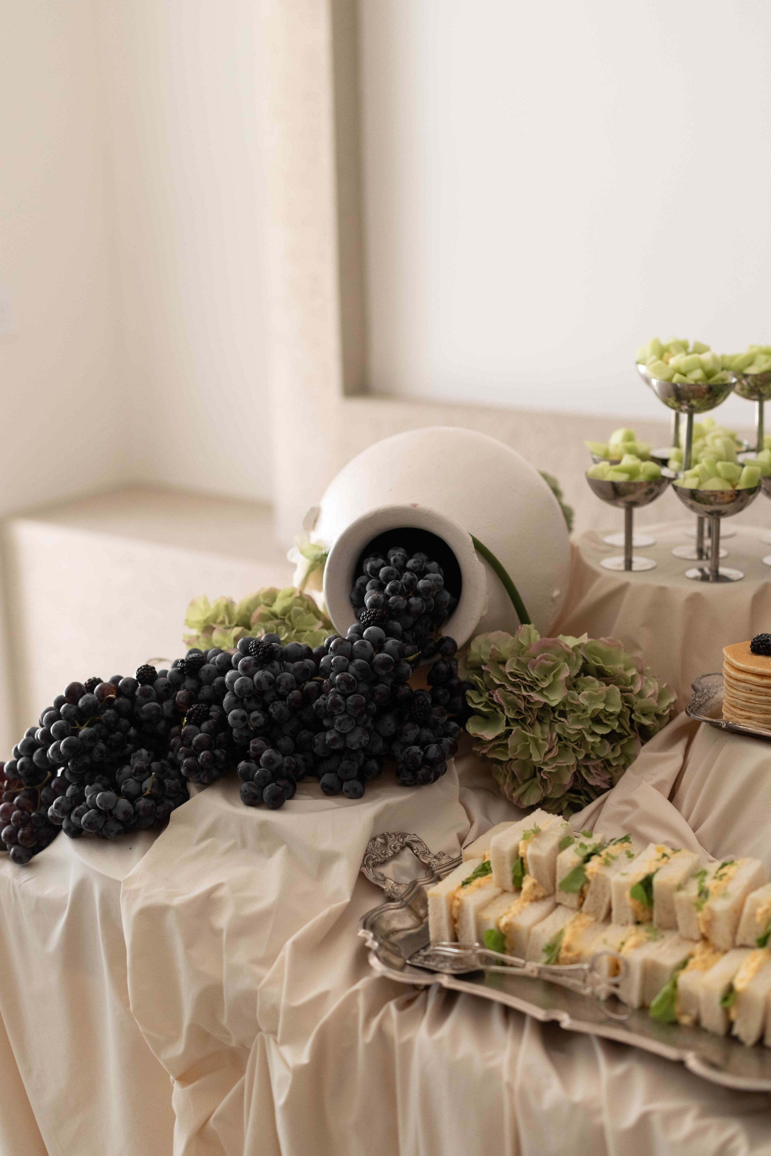 Elegant catering display with dark grapes spilling from white vase, finger sandwiches, and green salads on draped table.