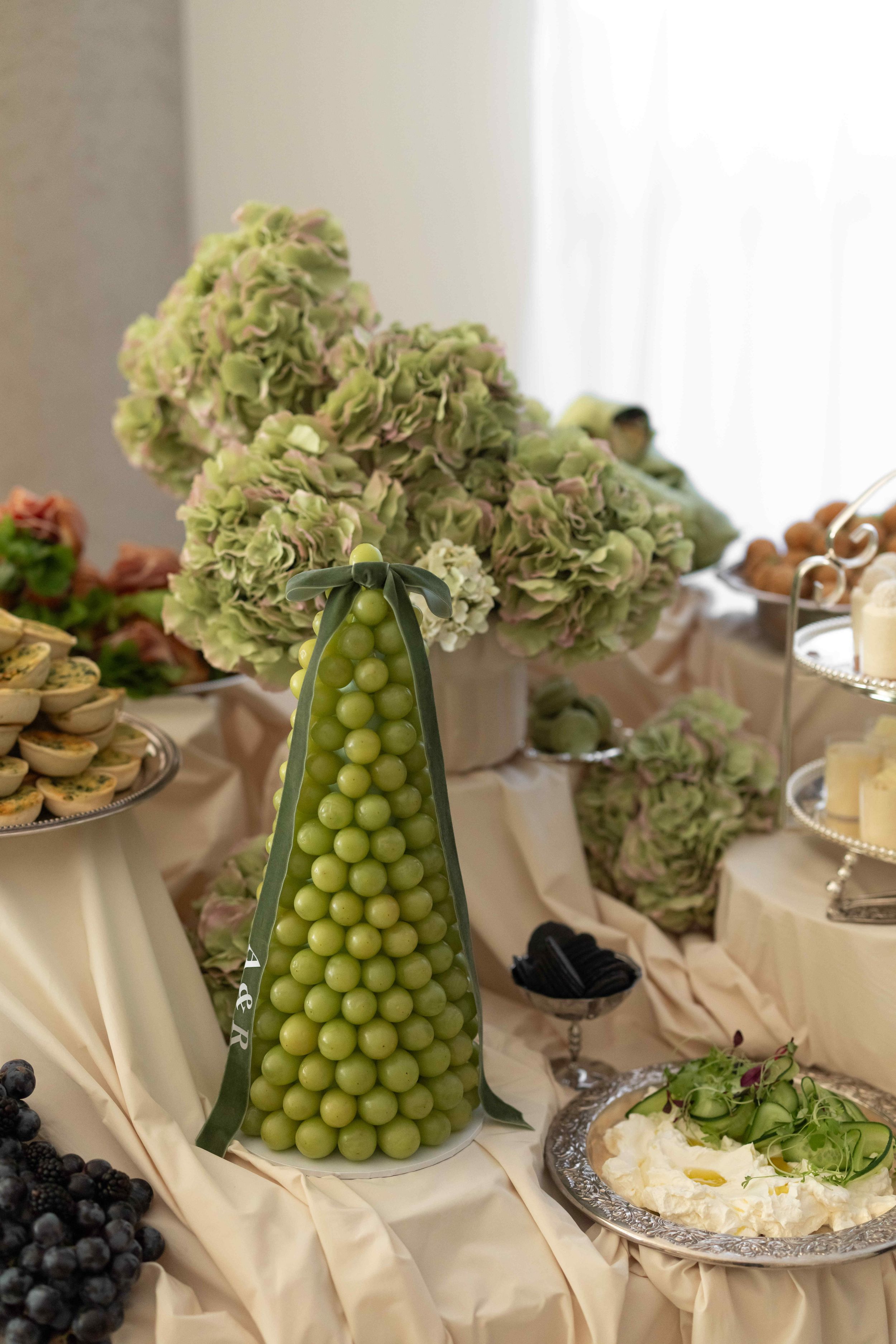 Elegant buffet table with green grape tower centerpiece, hydrangea flowers, cheese platter, and assorted appetizers.