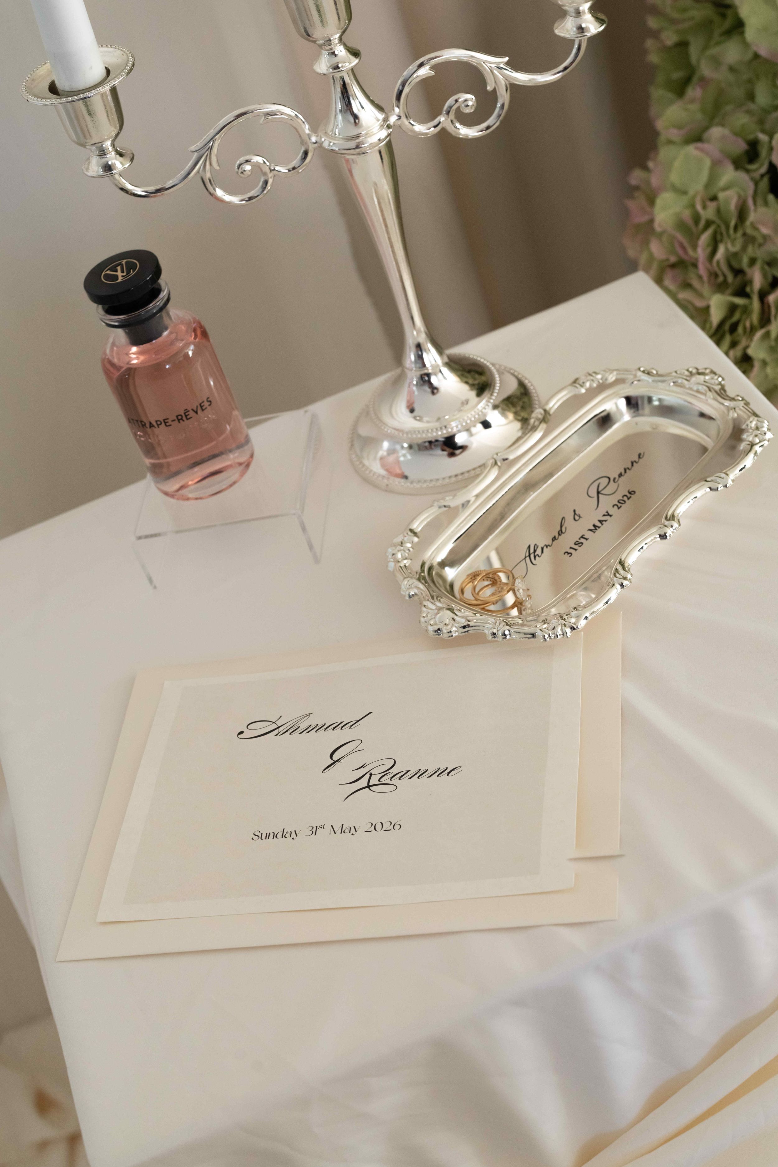 Elegant wedding stationery with calligraphy names on ivory card, silver candelabra, ring dish, and pink perfume bottle.