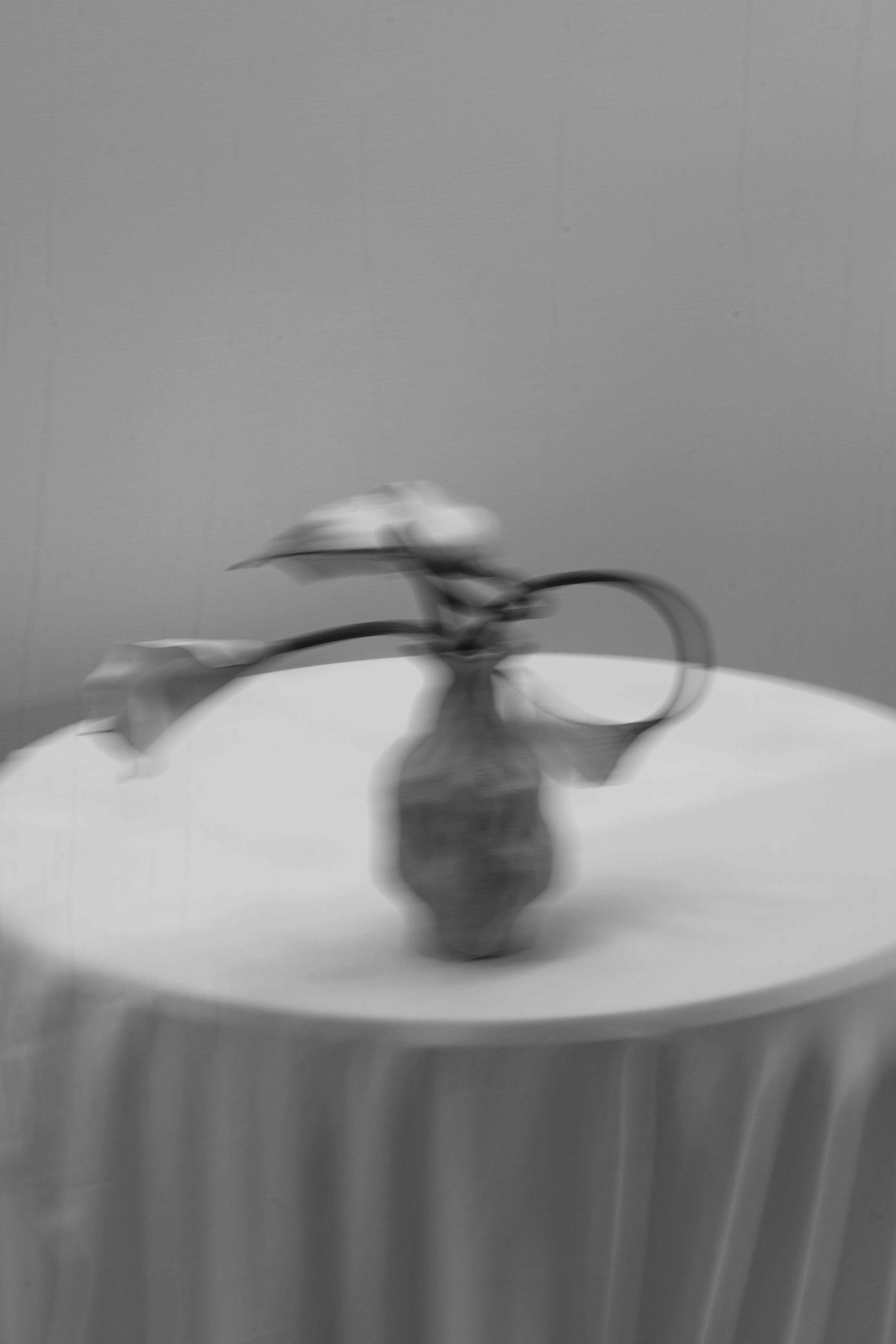 Blurry black and white photo of an ornate vase with flowers on a draped round table against a gray wall.