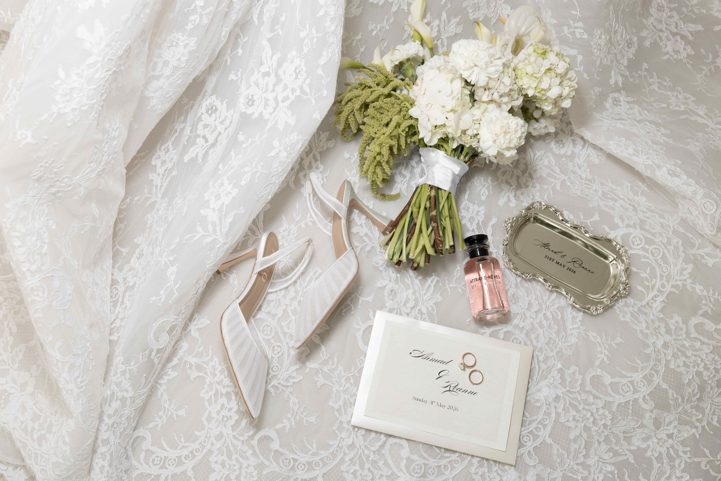 Elegant bridal flat lay with lace dress, nude heels, white floral bouquet, pink perfume, and wedding invitation.