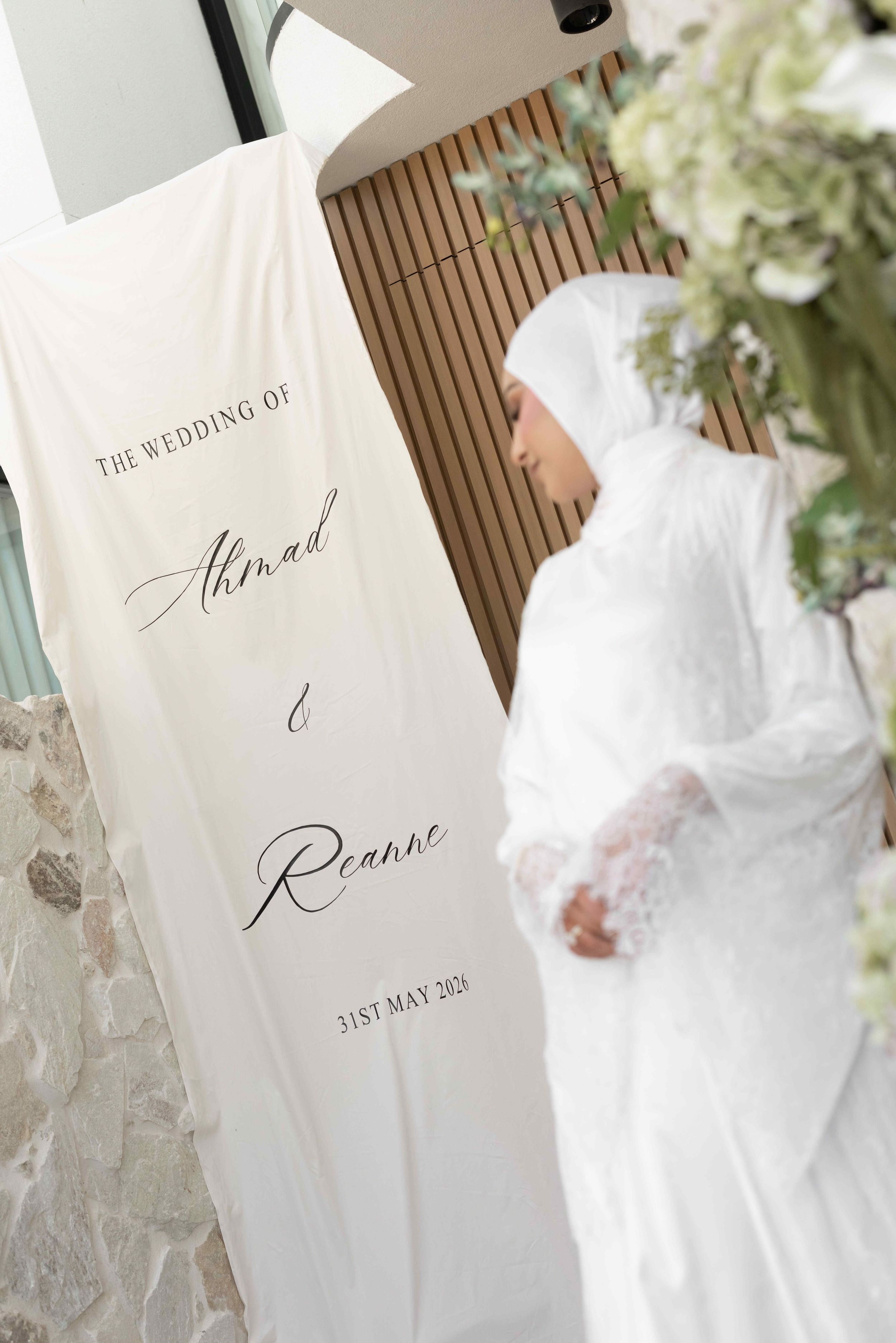 A bride in white hijab and lace gown stands beside a wedding banner reading Ahmad and Reanne, 31st May 2021.