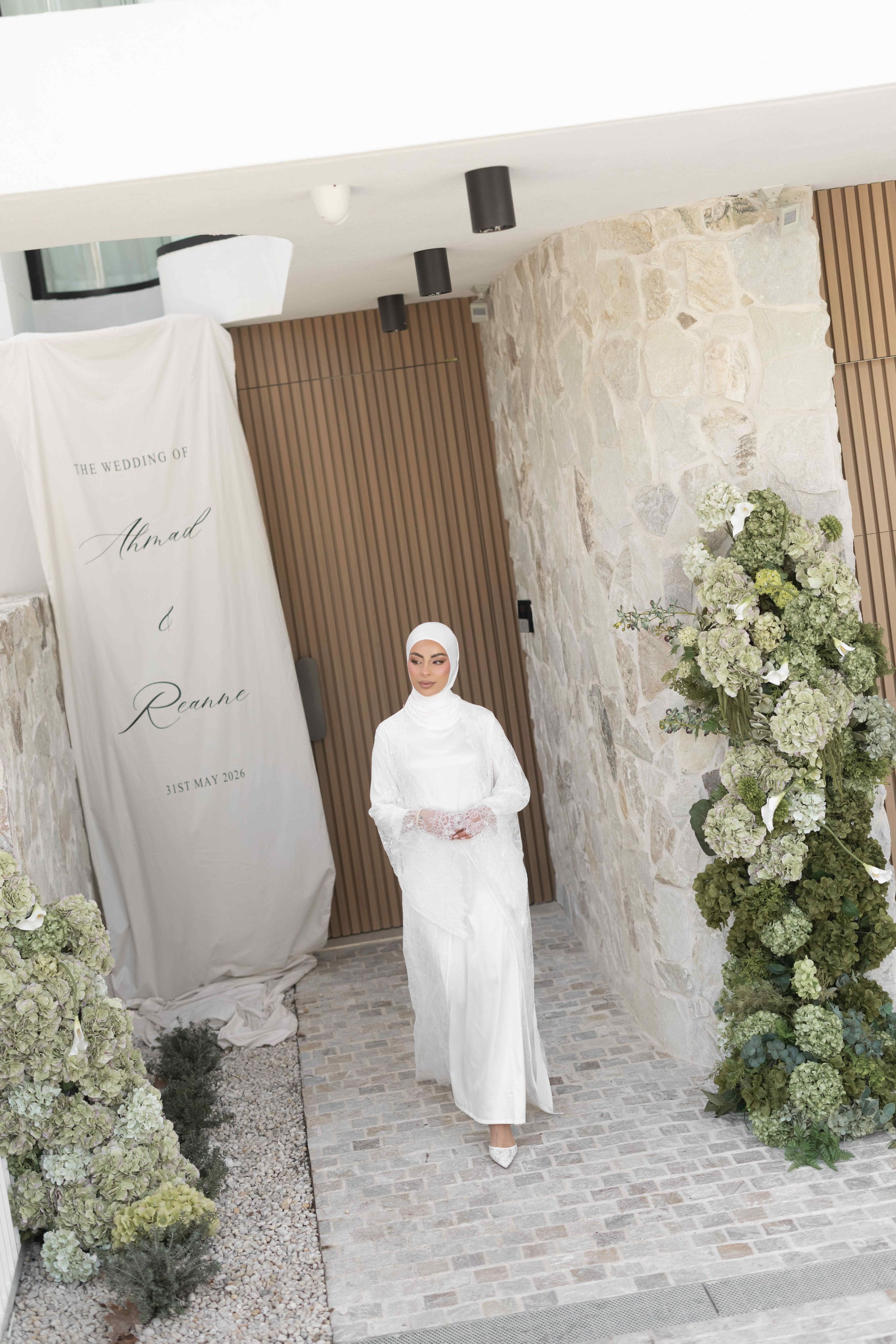 Bride in white hijab and gown stands at floral wedding entrance with hydrangeas and elegant banner.