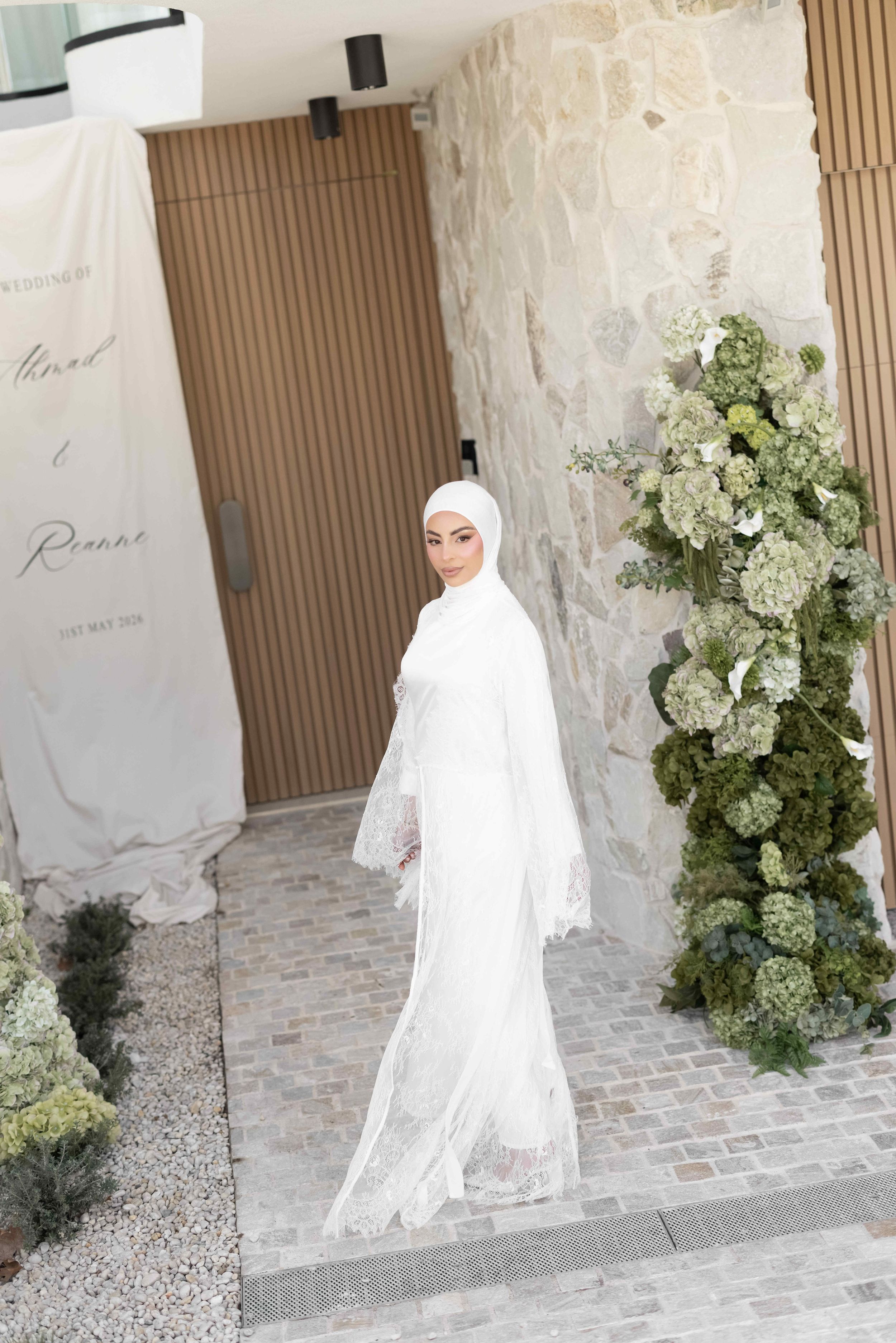Bride in white hijab and lace wedding gown standing by floral arch with hydrangeas at elegant venue.