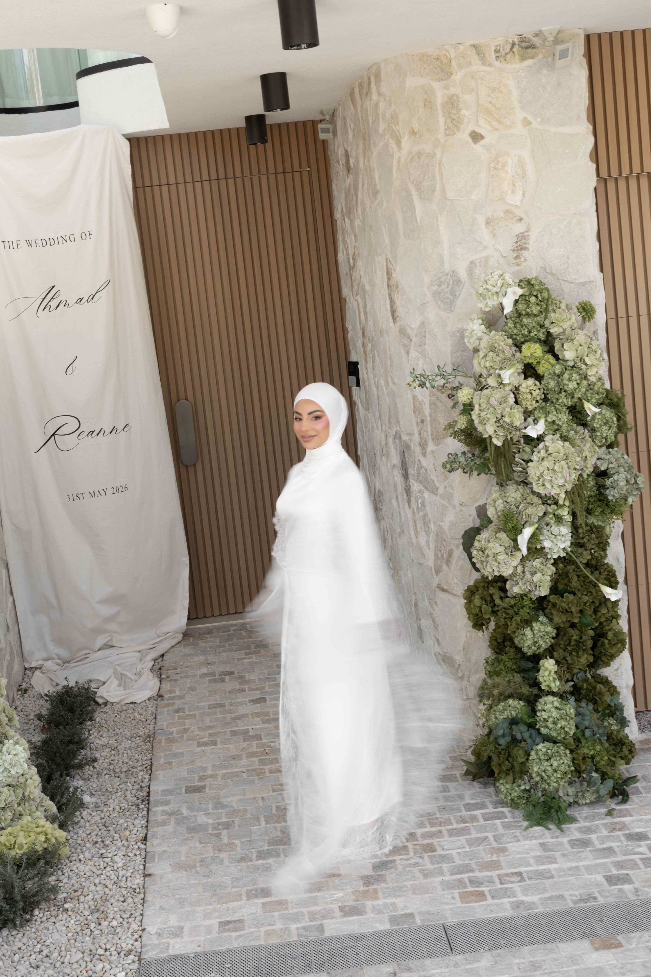 Bride in white hijab and wedding dress stands by floral arch at outdoor wedding venue entrance.