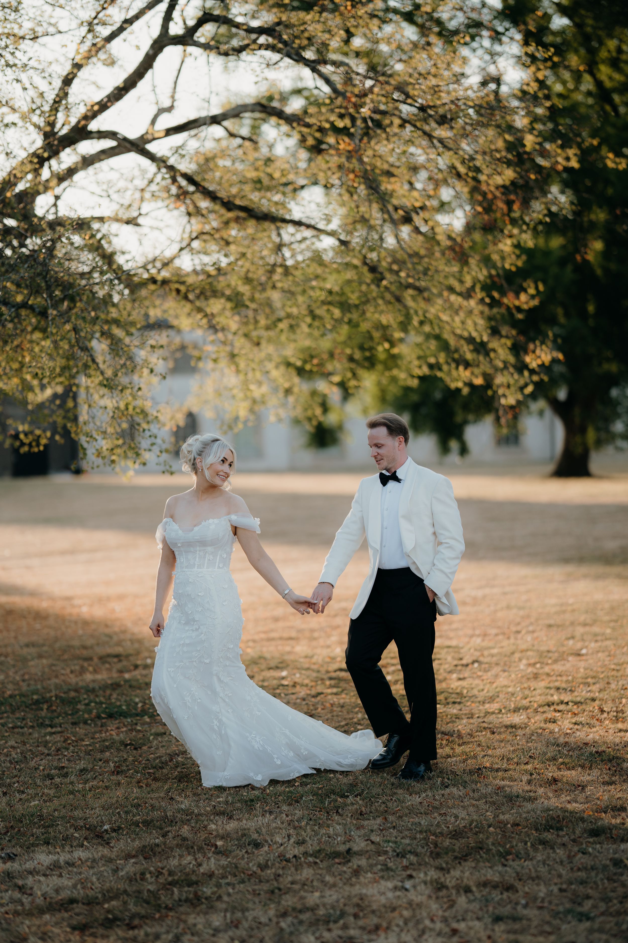 Bride and groom hold hands in golden hour light beneath autumn trees, she in lace gown, he in white tuxedo jacket.