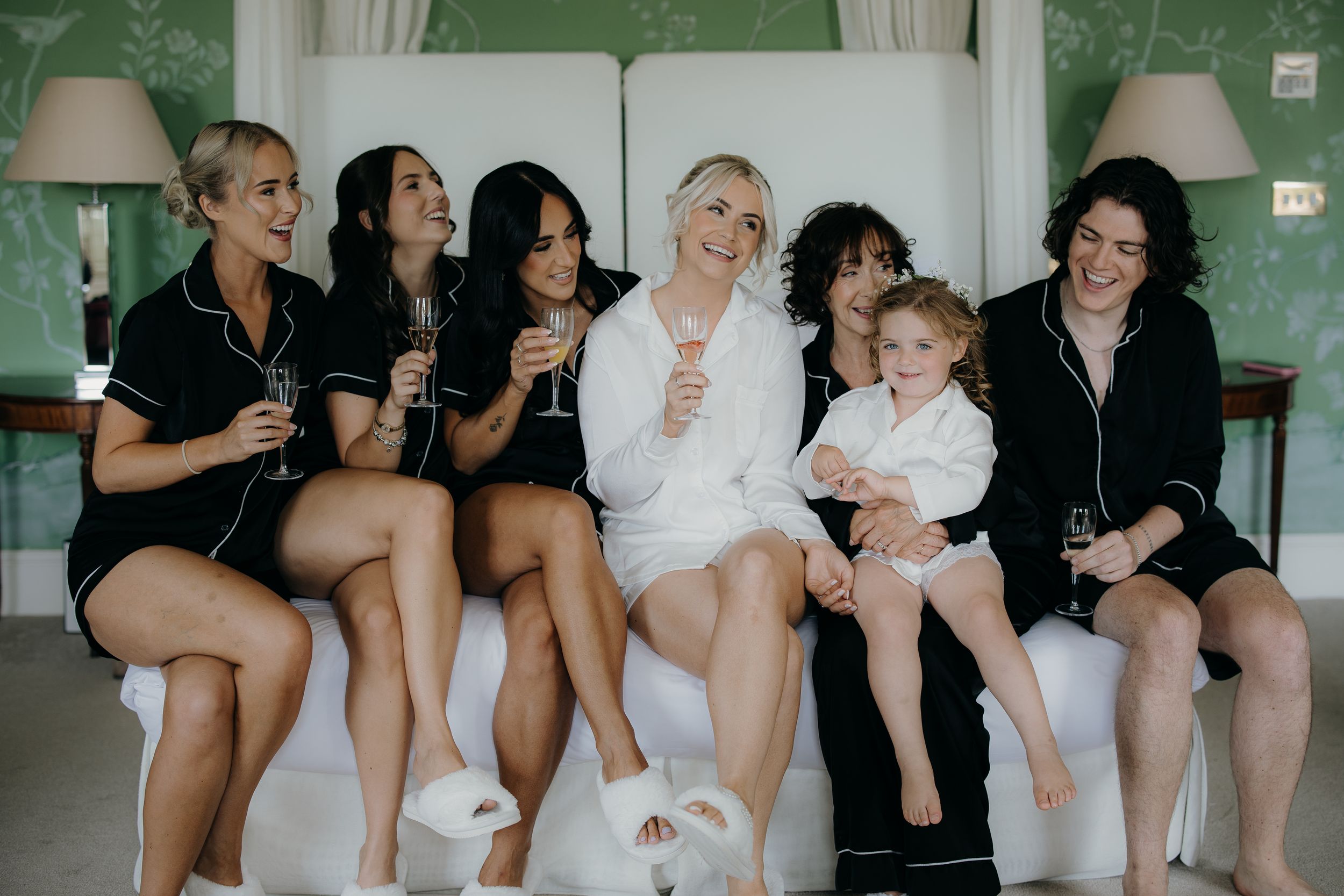 Bride in white robe with bridesmaids in black pajamas celebrating with champagne glasses in elegant green room.