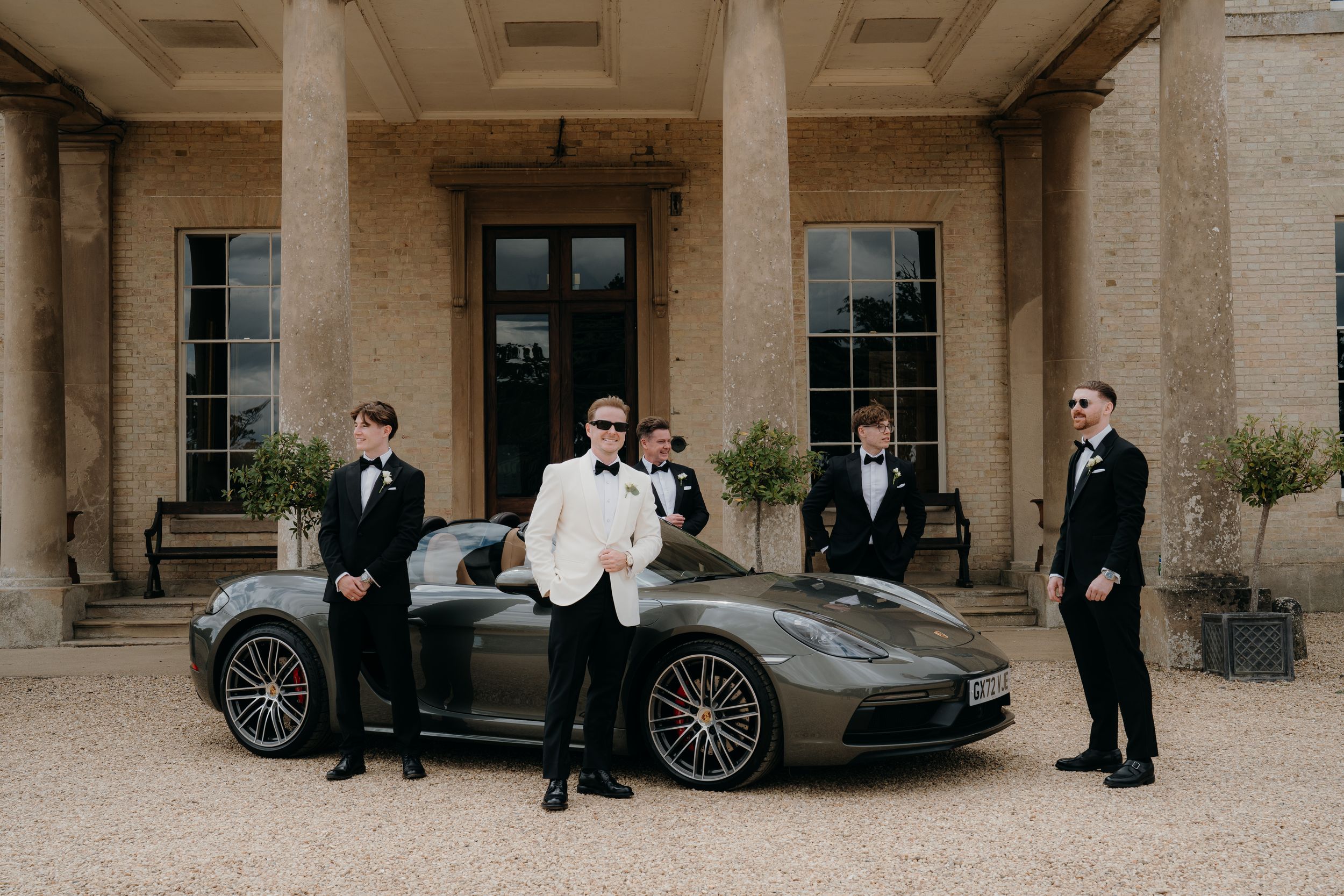 Groom in white tuxedo jacket poses with groomsmen in black suits beside a grey Porsche at an elegant estate.