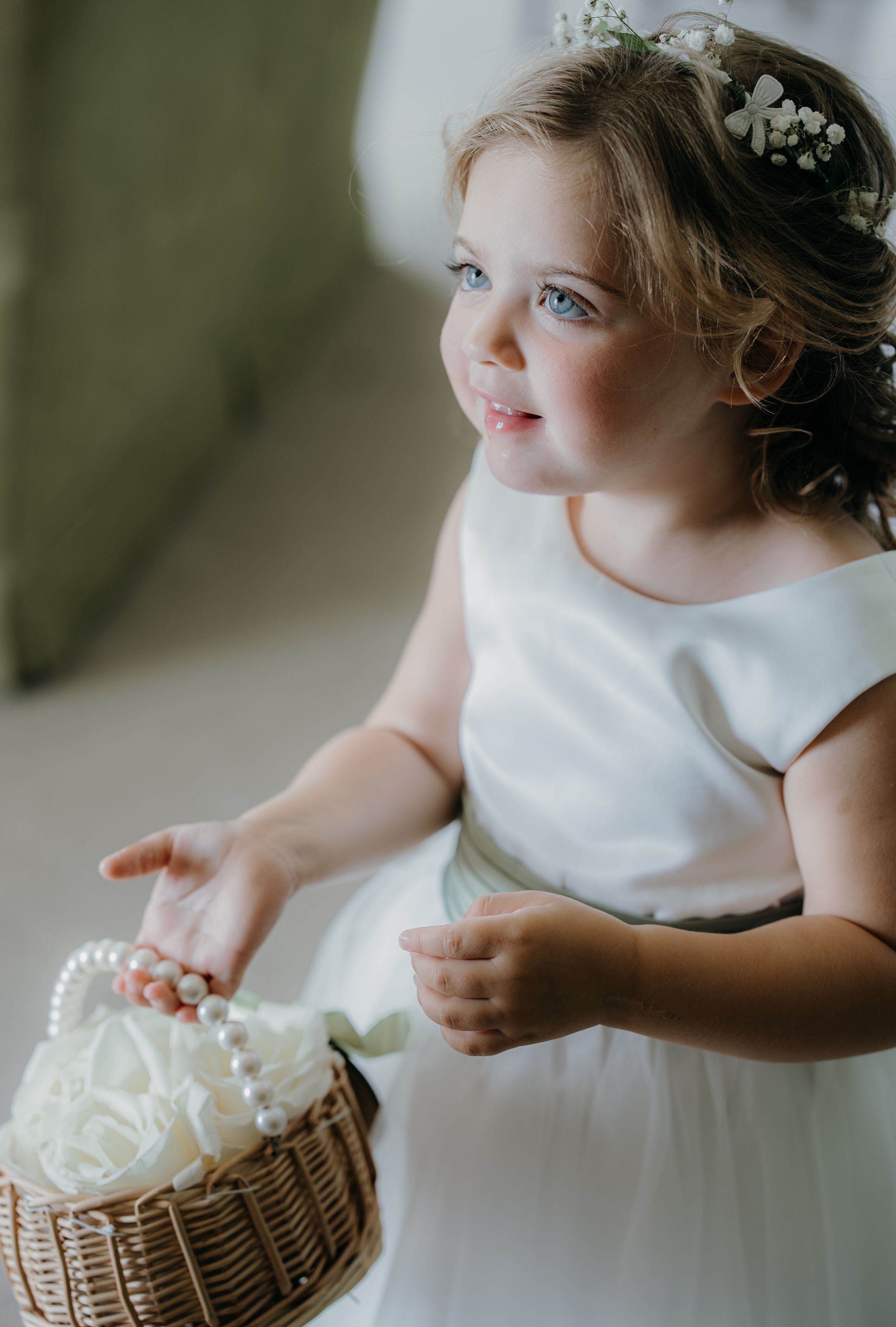 A young flower girl in a white dress holds a wicker basket with pearl handle and white roses, wearing a floral crown.