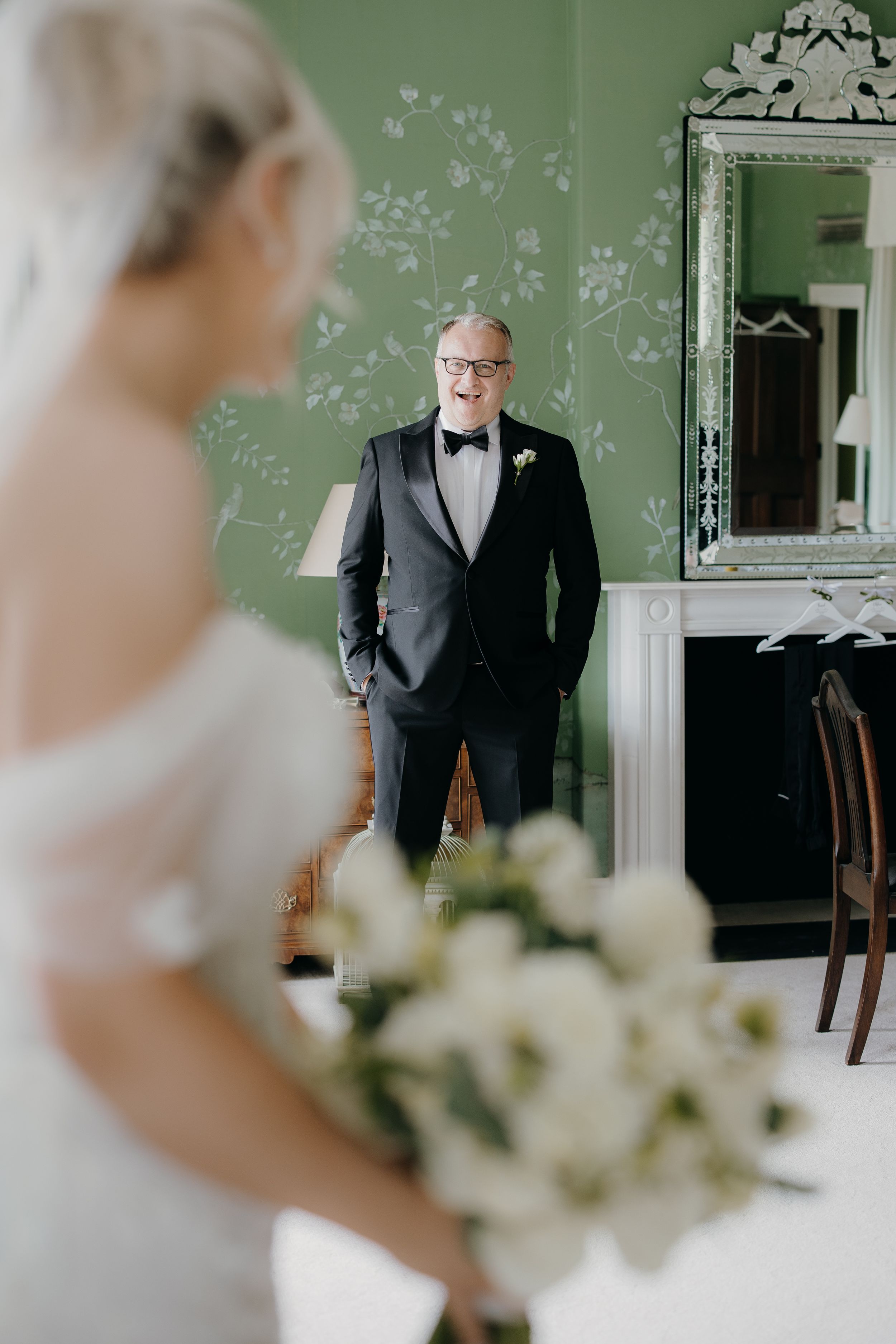 Father in tuxedo smiles at bride holding white bouquet during emotional first look in elegant green room.