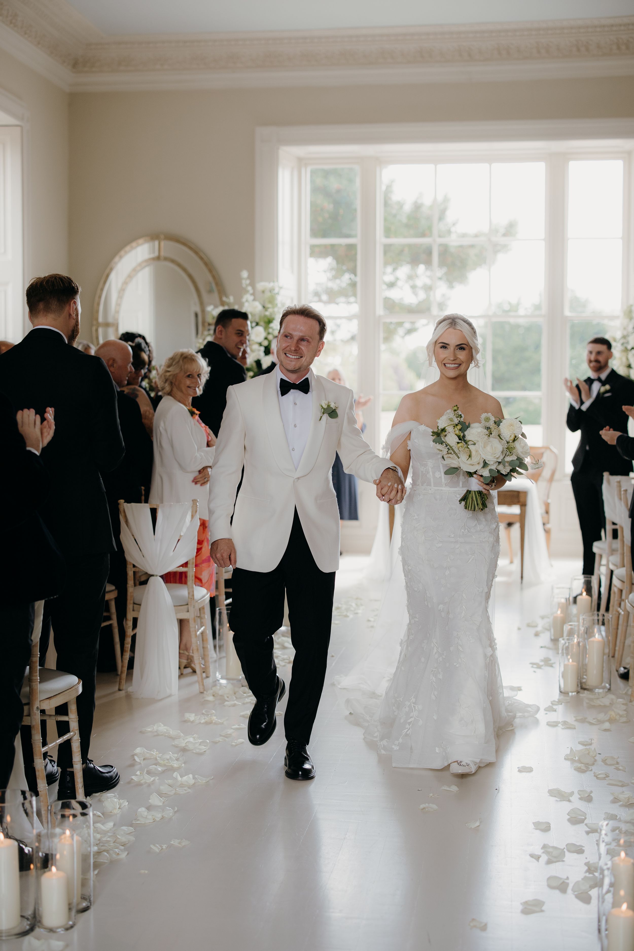 Bride and groom walk down the aisle hand in hand, smiling, in an elegant white ceremony room with rose petals.