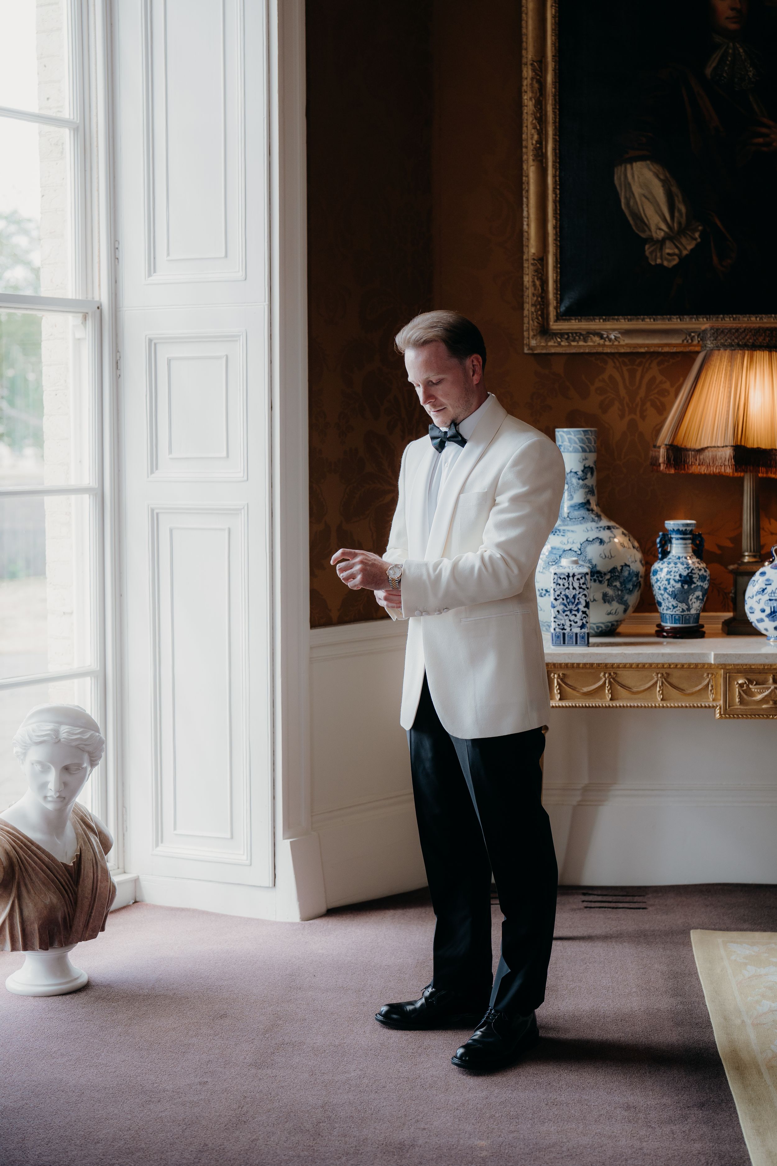 Groom in white tuxedo jacket adjusting cufflinks in elegant room with blue and white vases and classical bust.