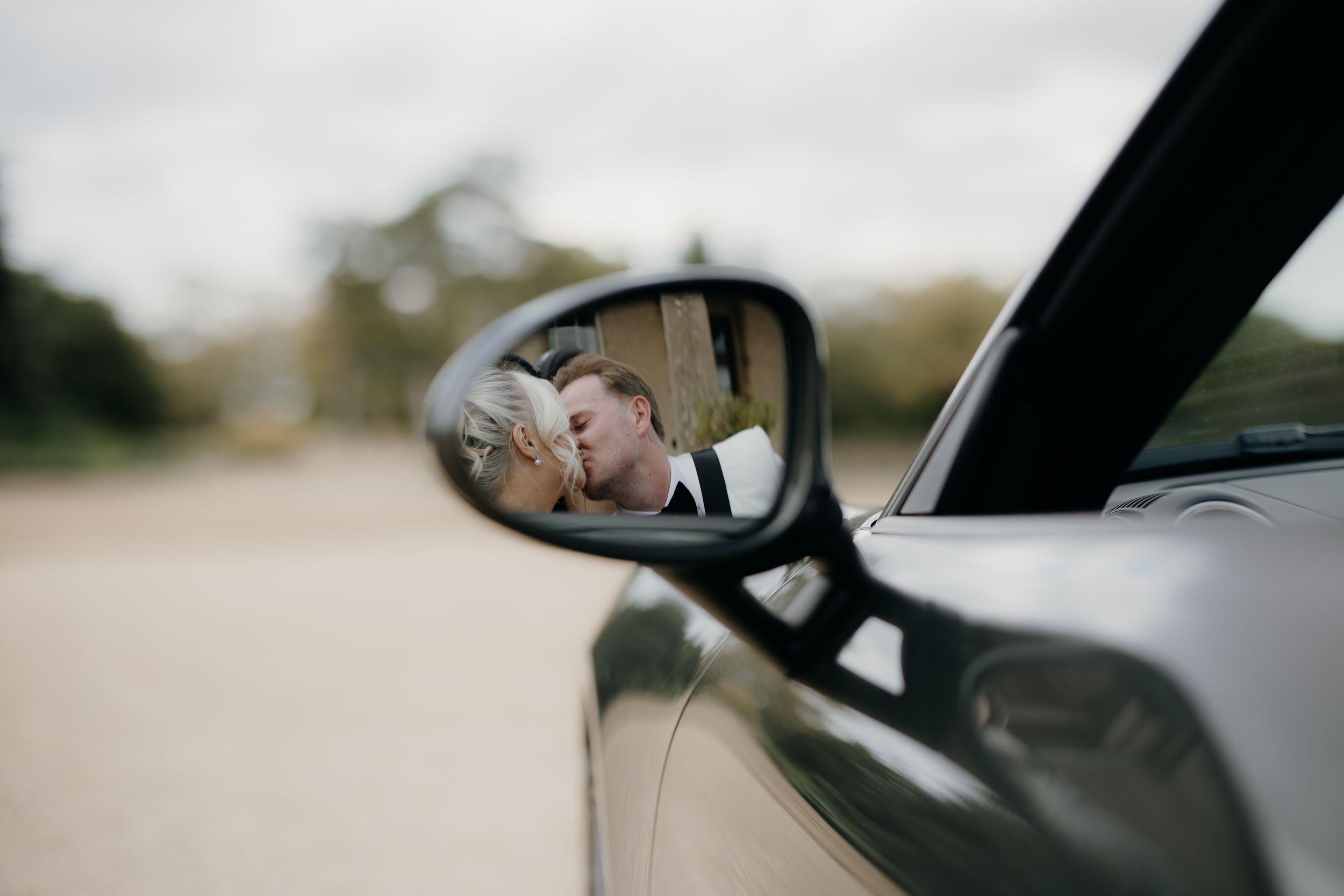 A groom in formal attire is reflected in a car side mirror, captured in an artistic wedding photography shot.