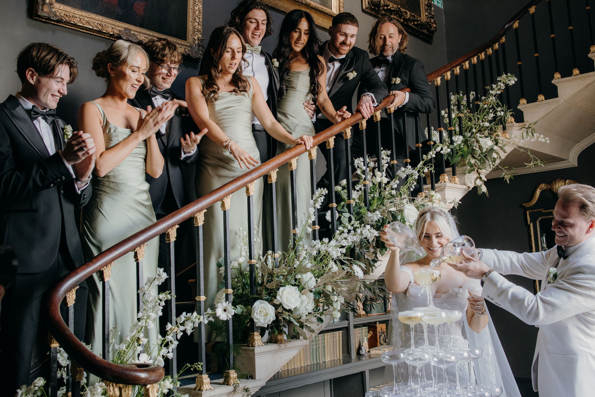 Wedding party on elegant staircase adorned with white floral arrangements, celebrating with a champagne tower.
