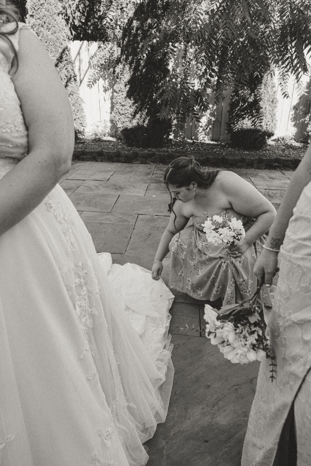 Bridesmaid in sequin dress bends to adjust bride's white gown train while holding floral bouquet at outdoor wedding.