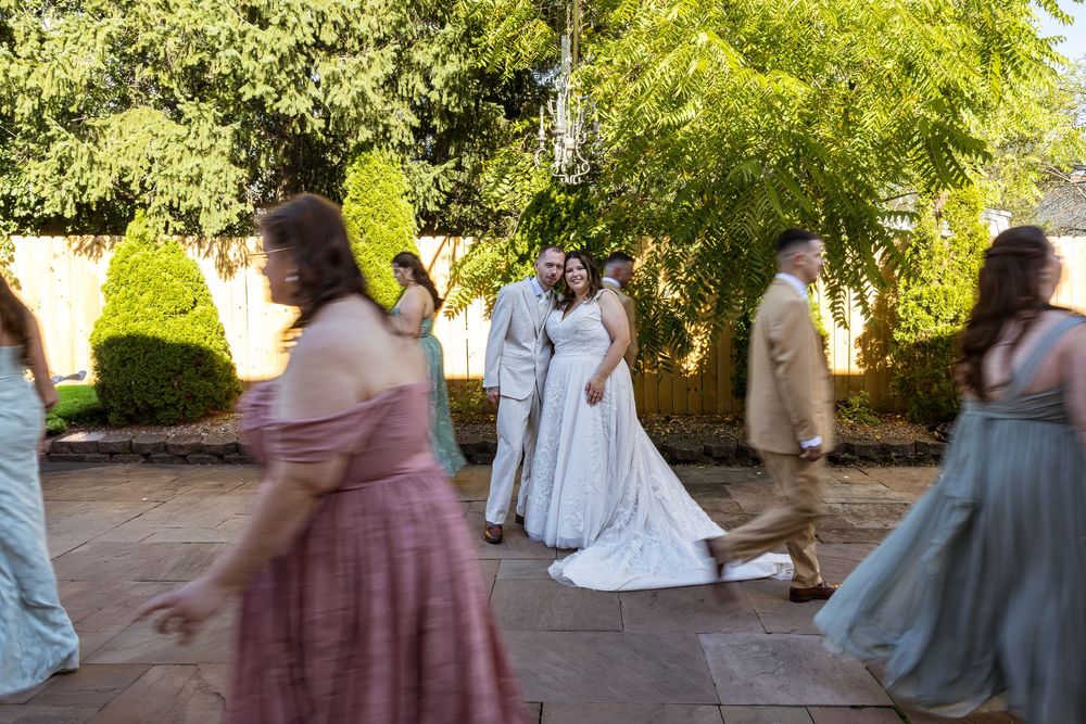 A bride and groom pose in a garden courtyard while wedding guests in colorful gowns move around them.