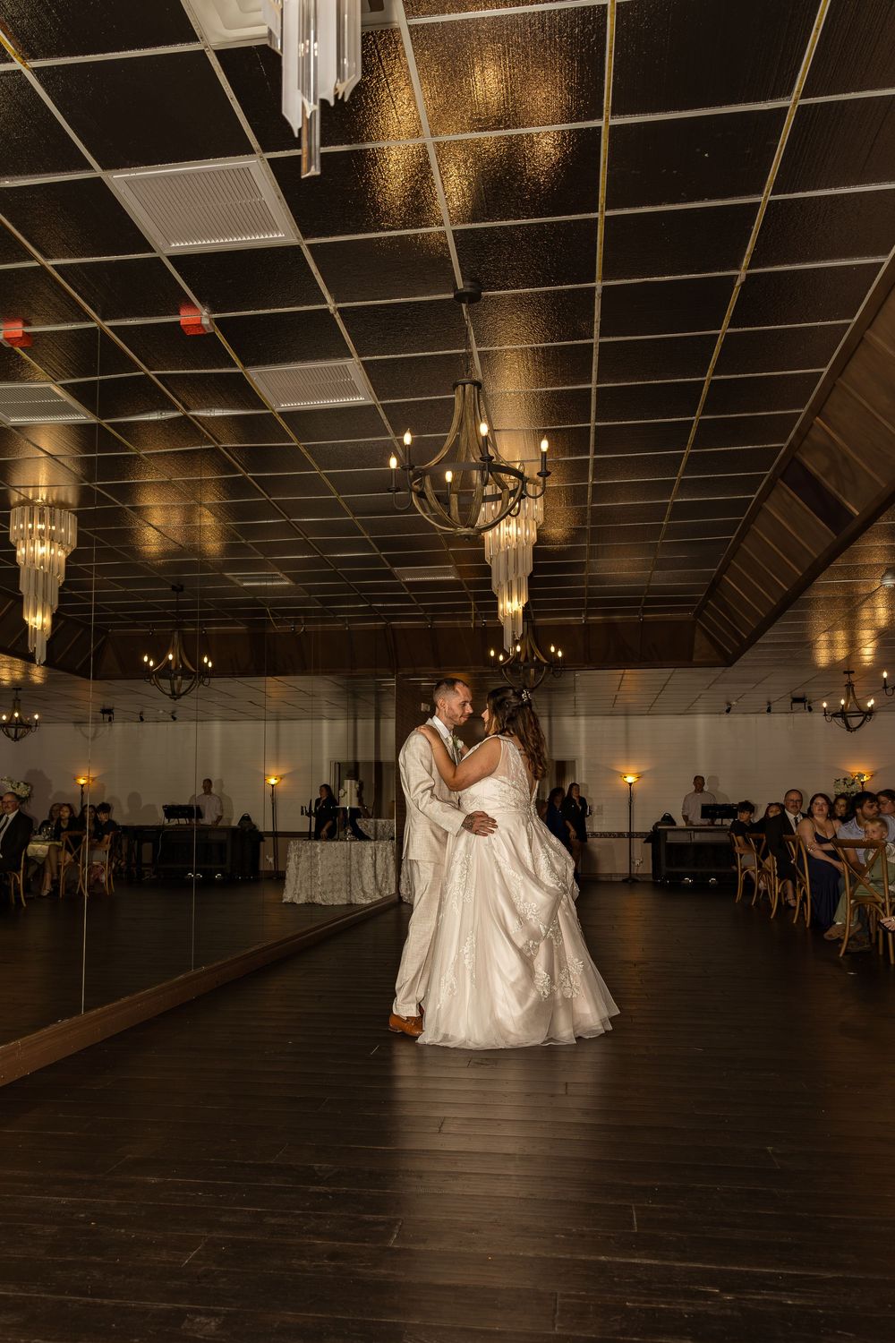 Bride and groom share their first dance on a dark hardwood floor beneath elegant chandeliers at a wedding reception.