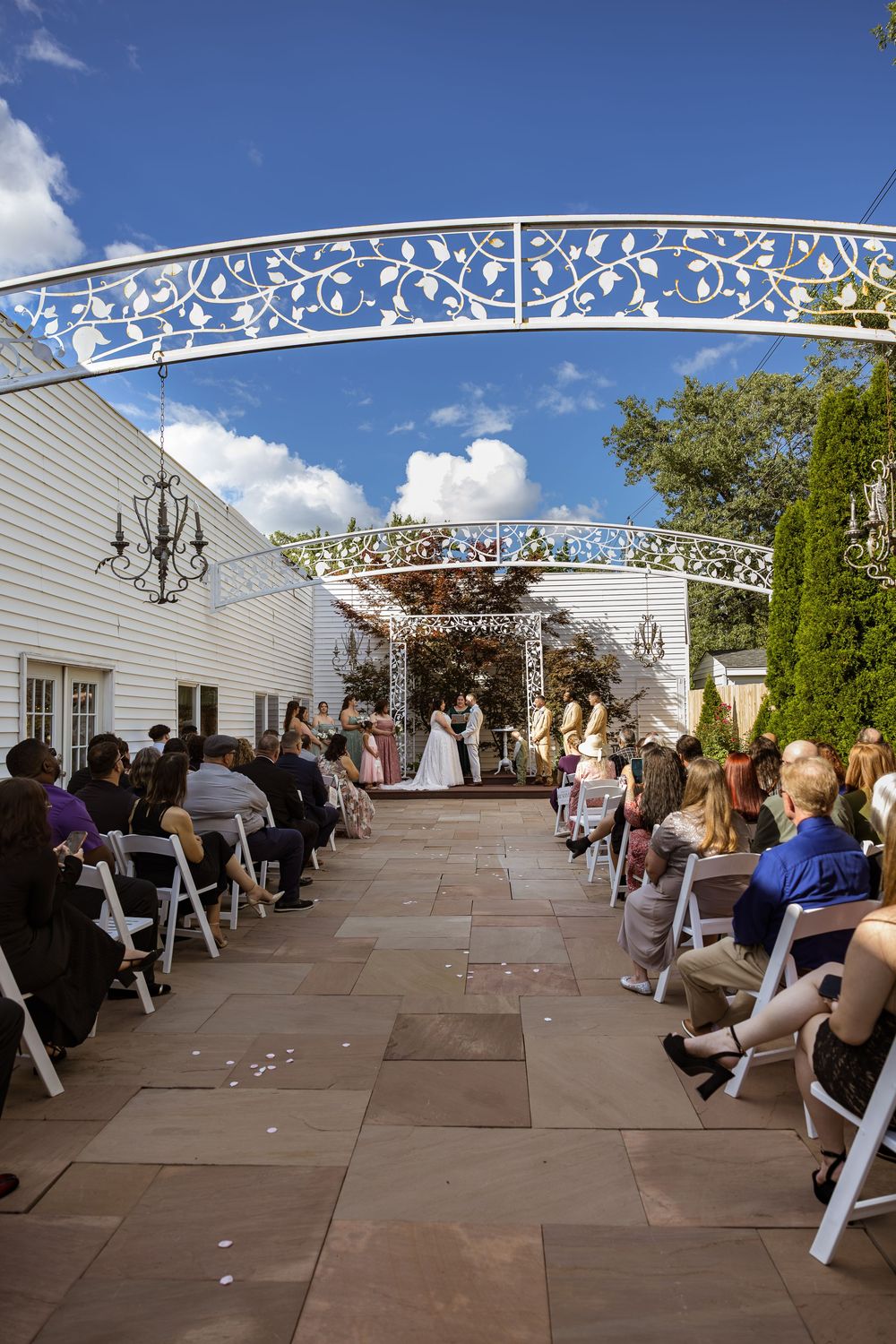 Outdoor wedding ceremony on a stone patio under decorative white arch with guests seated in white chairs on a sunny day.