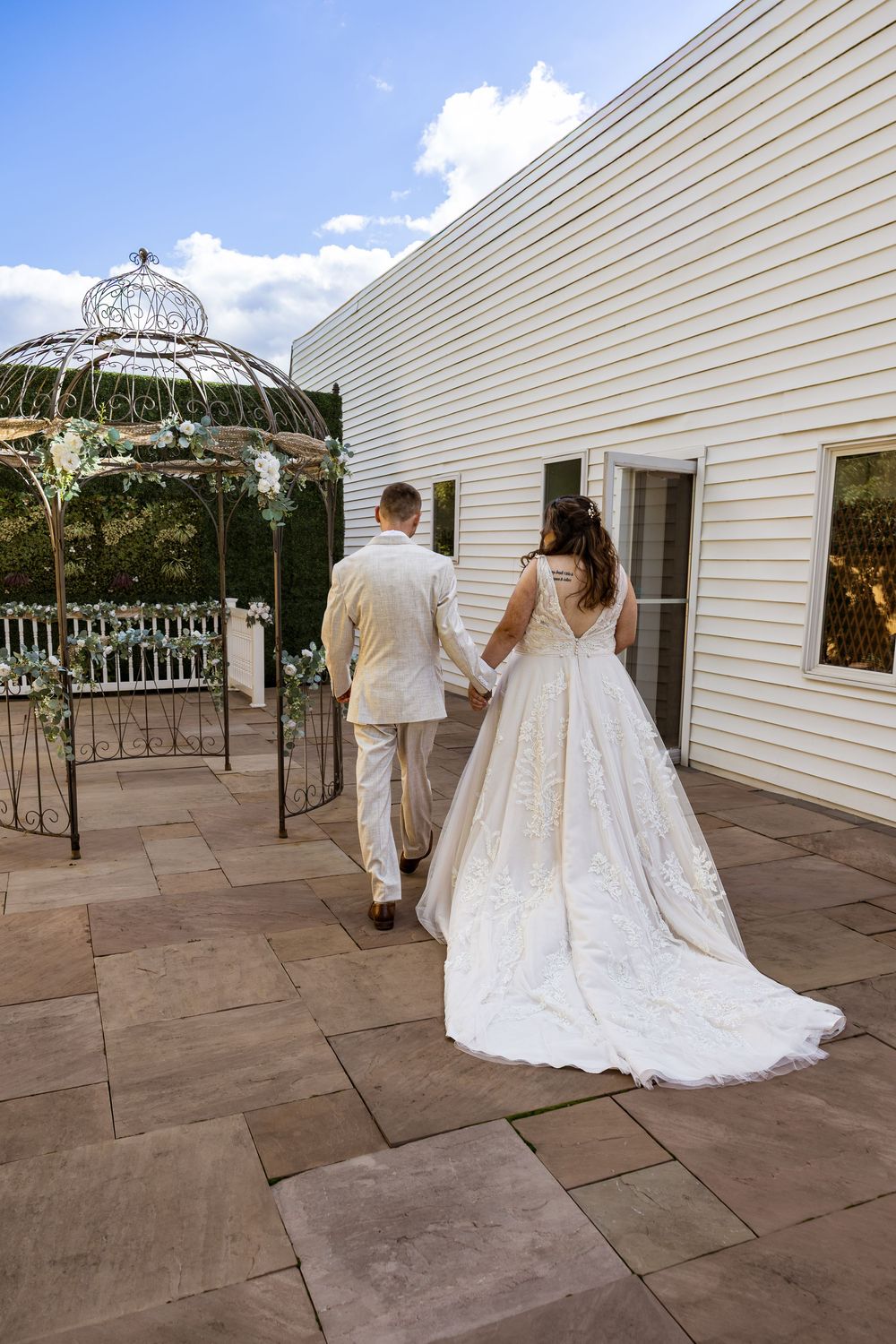 Bride in white gown and groom in tan suit walk hand-in-hand toward an elegant gazebo on a stone patio.